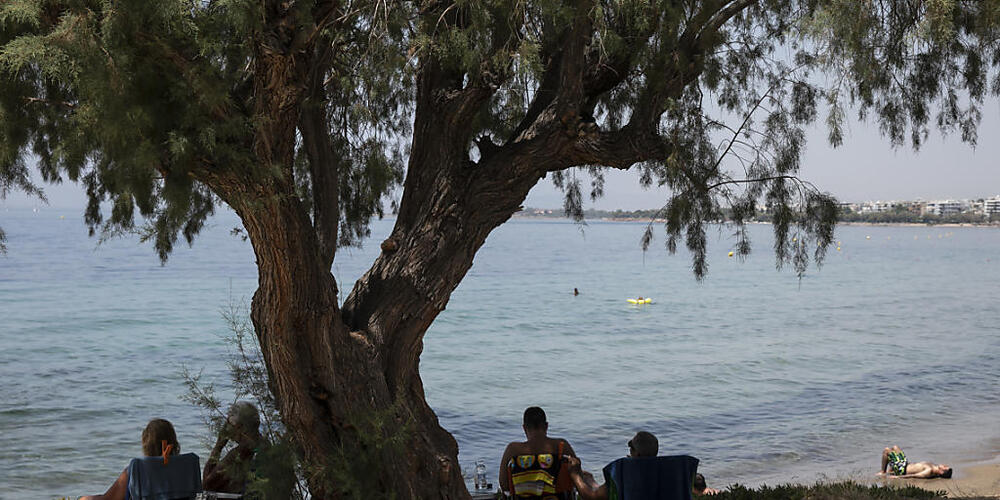Besucher sitzen im Schatten unter einem Pinienbaum am Strand des Vororts Kavouri südwestlich von Athen. Foto: Yorgos Karahalis/AP/dpa