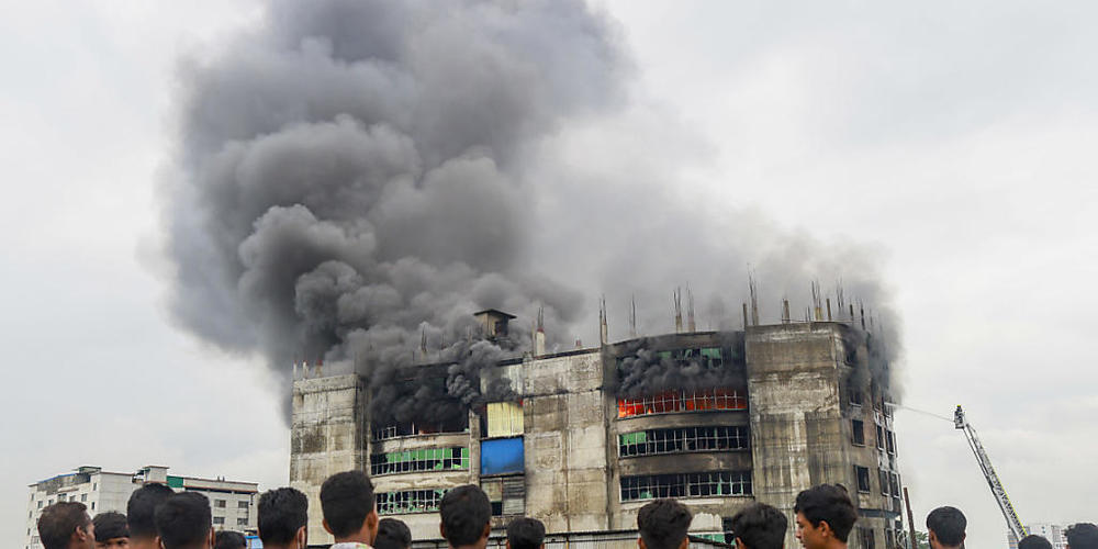 Menschen sehen zu, wie während des Brands in einer Fabrik Rauch von dem Gebäude aufsteigt. Bei einem Brand in einer Lebensmittel- und Getränkefabrik außerhalb der bangladeschischen Hauptstadt Dhaka sind mindestens 52 Menschen getötet worden. Foto:...