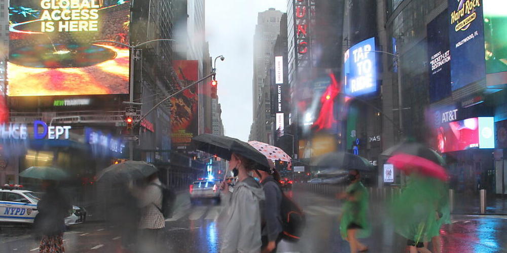 Fußgängerinnen und Fußgänger bei der Straßenüberquerung am Times Square schützen sich mit Regenschirmen vor starkem Regen. Der Tropensturm «Henri» hat in der U-Bahnstation Times Square - 42nd Street einen Wasserschaden verursacht. Foto: Niyi Fote/...