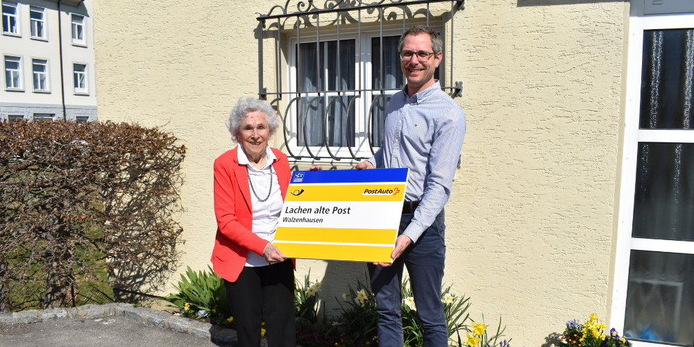 Michael Litscher übergibt Heidi Sturzenegger-Knellwolf die geschichtsträchtige PostAuto-Haltestelle-Tafel „Lachen alte Post“ 