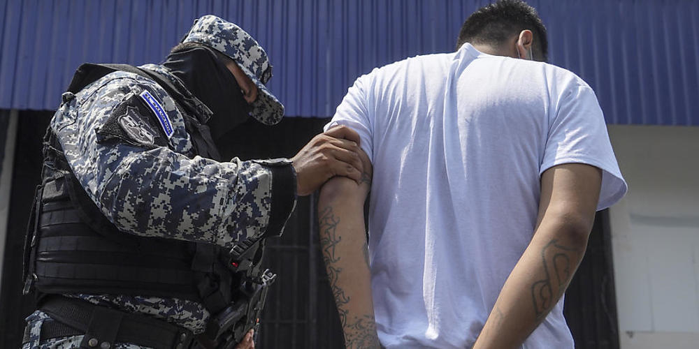 ARCHIV - Ein vermummter Polizist begleitet ein mutmaßliches Bandenmitglied nach dessen Festnahme. Während des seit gut einem Monat andauernden Ausnahmezustands sind in El Salvador mehr als 20 000 mutmaßliche Bandenmitglieder festgenommen worden. D...
