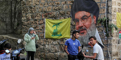 Menschen sitzen in einer Straße mit dem Bild des Hisbollah-Führers Nasrallah und Fahnen, die während der libanesischen Parlamentswahlen 2022 in der Nähe eines Wahllokals in Beirut aufgestellt wurden. Die Libanesen haben inmitten der schwersten Wir...