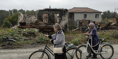 Zwei Frauen gehen mit ihren Fahrrädern auf einer Straße neben zerstörten russischen Schützenpanzer und zerstörten Häusern spazieren. Foto: Evgeniy Maloletka/AP/dpa