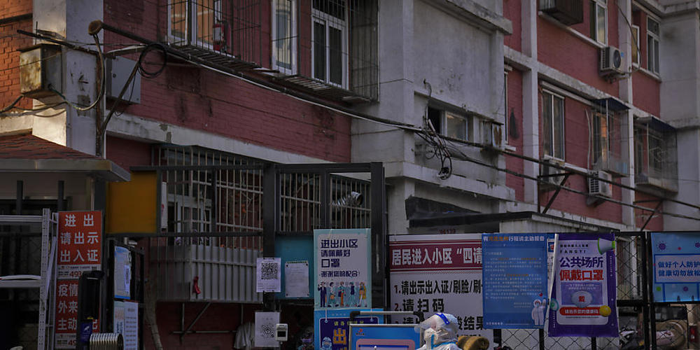 Ein Wachmann im Schutzanzug sitzt an einem Eingangstor zu einem Wohnviertel in Peking. Vor dem Hintergrund steigender Corona-Zahlen haben zwei der größten Städte in China am 21.11.2022 weitere Einschränkungen angekündigt. Foto: Andy Wong/AP/dpa
