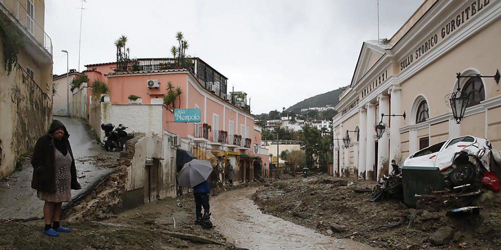 Menschen stehen an einer mit Schlamm und Wasser bedeckten Straße, nachdem schwere Regenfälle Erdrutsche ausgelöst hatten. Auf der italienischen Insel Ischia sind nach einem schweren Unwetter und Erdrutschen mehrere Menschen als vermisst gemeldet w...