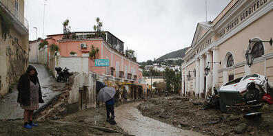 Menschen stehen an einer mit Schlamm und Wasser bedeckten Straße, nachdem schwere Regenfälle Erdrutsche ausgelöst hatten. Auf der italienischen Insel Ischia sind nach einem schweren Unwetter und Erdrutschen mehrere Menschen als vermisst gemeldet w...