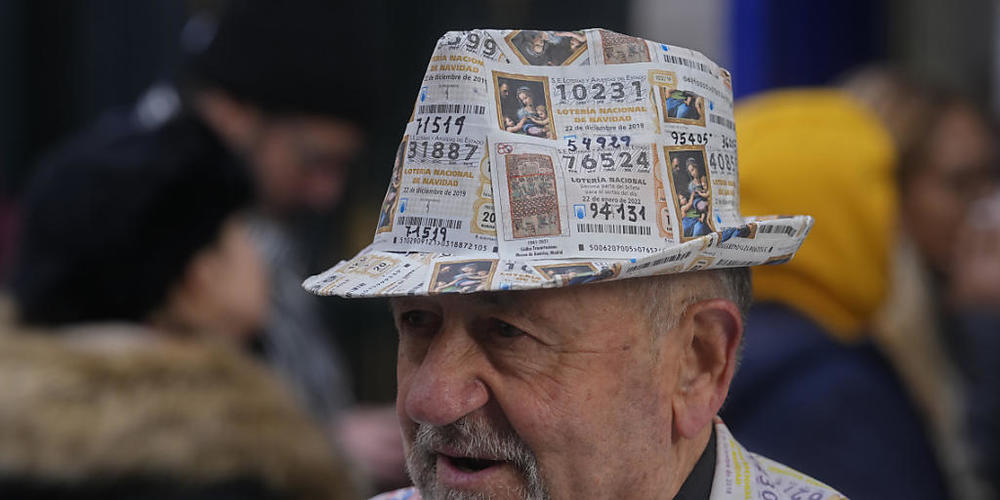 Ein Mann mit einem Anzug und Hut, der mit gedruckten Lottoscheinen verziert ist, steht vor dem berühmten Lotterieladen «Dona Manolita» in der Innenstadt. Foto: Paul White/AP/dpa