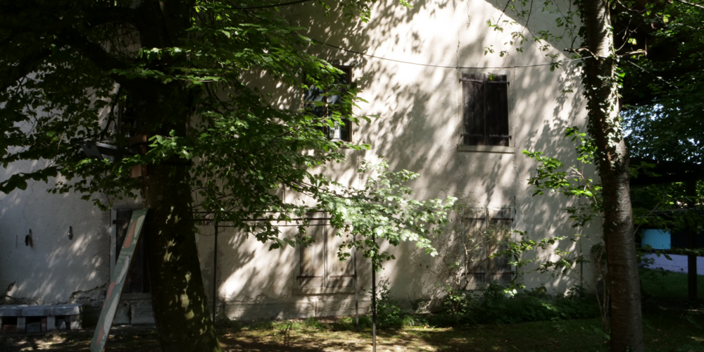 Von Bäumen partiell verdeckte Fassade des Baumwollmagazins der ehemaligen Spinnerei Schönau.