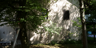 Von Bäumen partiell verdeckte Fassade des Baumwollmagazins der ehemaligen Spinnerei Schönau.