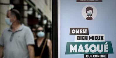 Passanten sind hinter einem Plakat mit der Aufschrift «On est bien mieux masqué que confiné» (Lieber maskiert als eingesperrt) zu sehen. Foto: Philippe Lopez/AFP/dpa