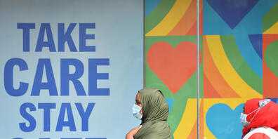 Frauen mit Kopftuch und Mundschutz gehen in einem Stadtzentrum in Großbritannien an einem Plakat vorbei, das an die Corona-Schutzregeln erinnert: "Take care, stay safe". Foto: Jacob King/PA Wire/dpa