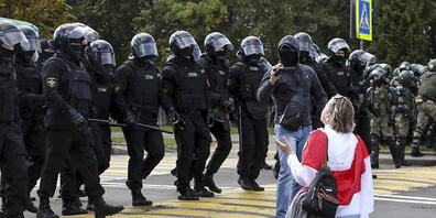 Eine Demonstrantin, die sich eine historische Flagge von Belarus um die Schultern gehängt hat, kniet vor einer Reihe von Polizisten in schwerer Montur auf dem Boden. Foto: -/TUT.by via AP/dpa