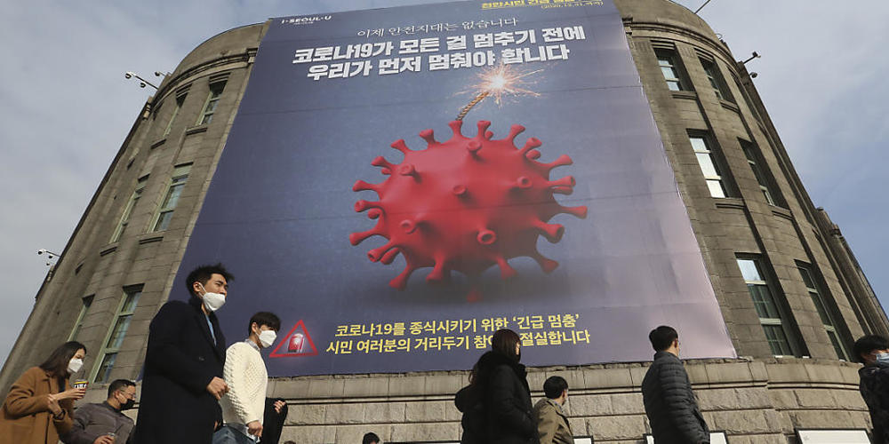 Menschen, die Gesichtsmasken tragen, gehen vor dem Rathaus von Seoul unter einem Transparent, das auf eine verstärkte soziale Distanzierungskampagne hinweist vorbei. Foto: Ahn Young-Joon/AP/dpa
