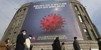 Menschen, die Gesichtsmasken tragen, gehen vor dem Rathaus von Seoul unter einem Transparent, das auf eine verstärkte soziale Distanzierungskampagne hinweist vorbei. Foto: Ahn Young-Joon/AP/dpa