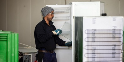 Ein Mitarbeiter eines Recyclingbetriebs räumt lose Teile aus einem ausgedienten Kühlschrank, bevor dieser durch einen zweitstufigen, maschinellen Prozess verarbeitet wird. 