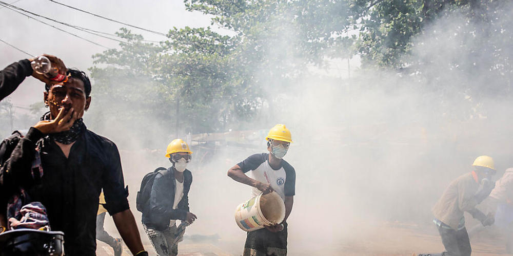 Gegner der Militärjunta von Myanmar laufen bei einem Protest gegen die Junta über die Straße und kippen Wasser auf rauchende Tränengaskanister. Die neue Junta in Myanmar geht weiter mit brutaler Gewalt und Inhaftierungen gegen politische Gegner, D...