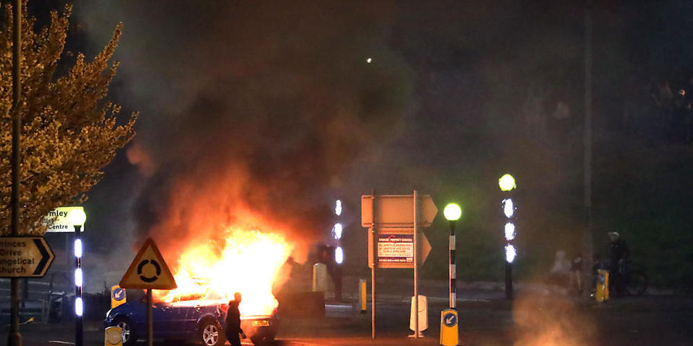 Ein Mann geht an einem brennenden Auto vorbei. Bei Protesten im nordirischen Belfast sind mehrere Polizisten verletzt worden. Foto: Peter Morrison/PA Wire/dpa